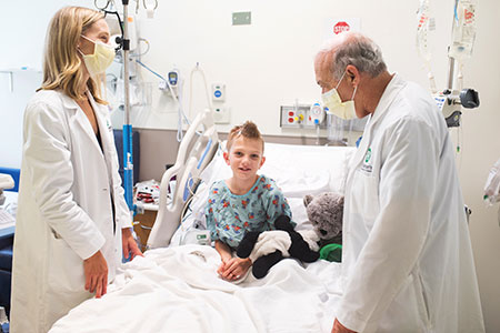 child in hospital bed with two caregivers standing at the side